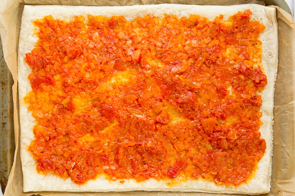 Overhead shot of puff pastry with spread out tomato sauce on a baking tray