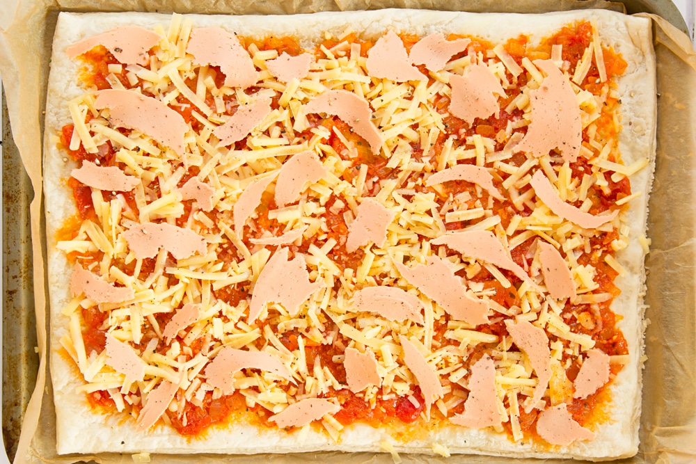 Overhead shot of puff pastry and tomato sauce topped with cheese and ham in a baking tray