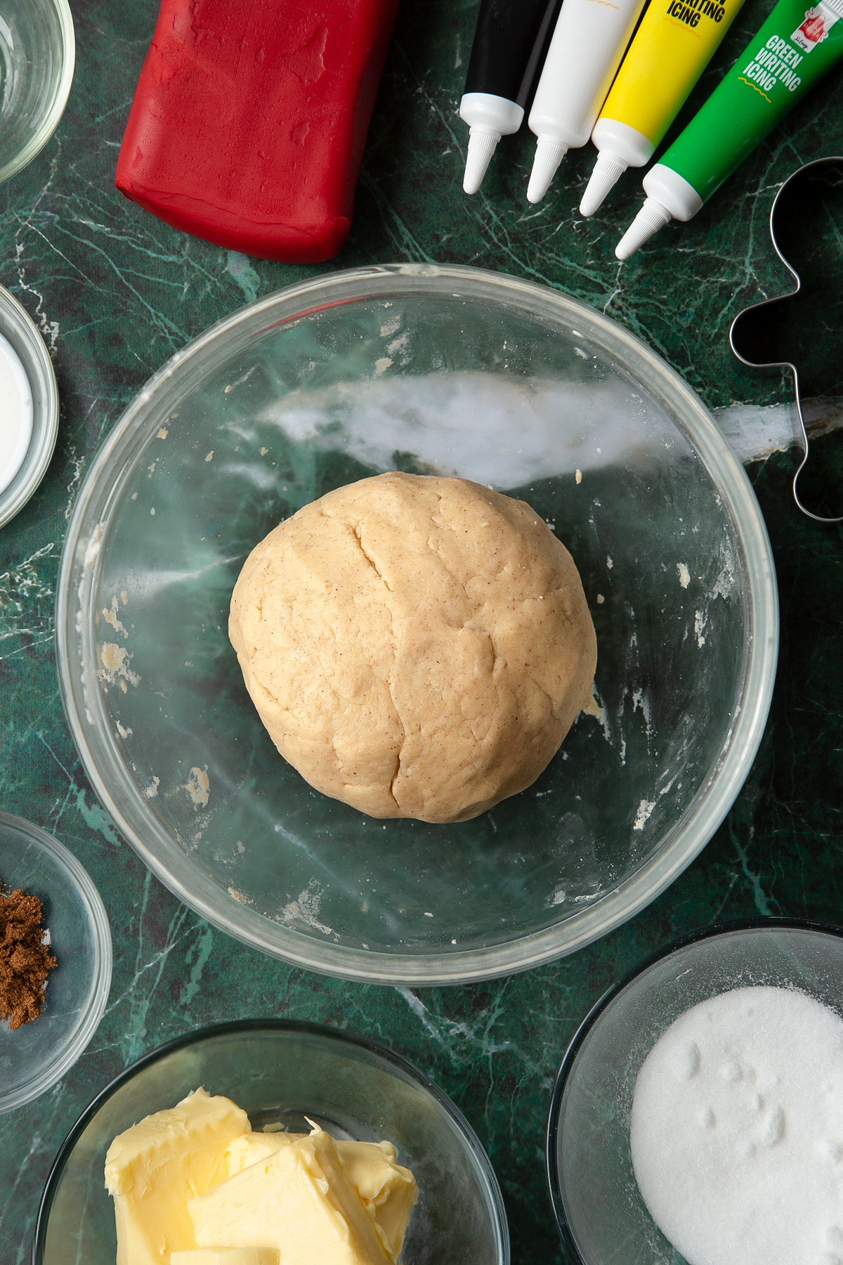 A ball of cookie dough in a glass mixing bowl. The bowl is surrounded by ingredients to make Father Christmas cookies.