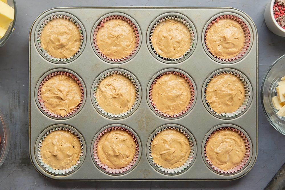 A twelve hole muffin tray lined with muffin cases and the spiced Christmas cupcakes with marzipan frosting cake mixture.