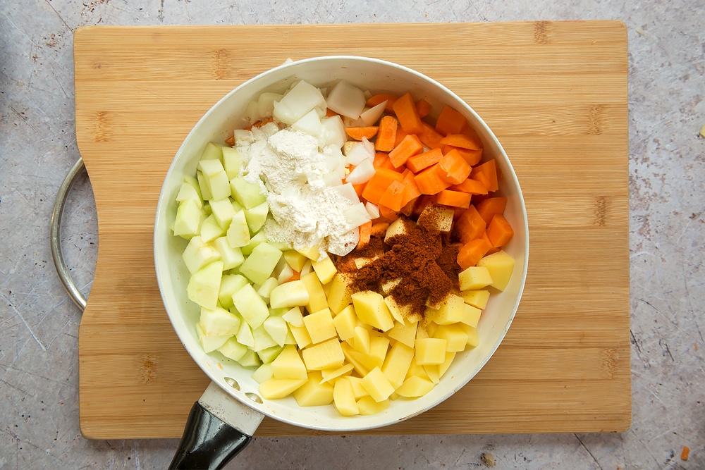 Overhead shot of sultanas, onion, carrots, apple, potato, curry powder and flour in a large white pan