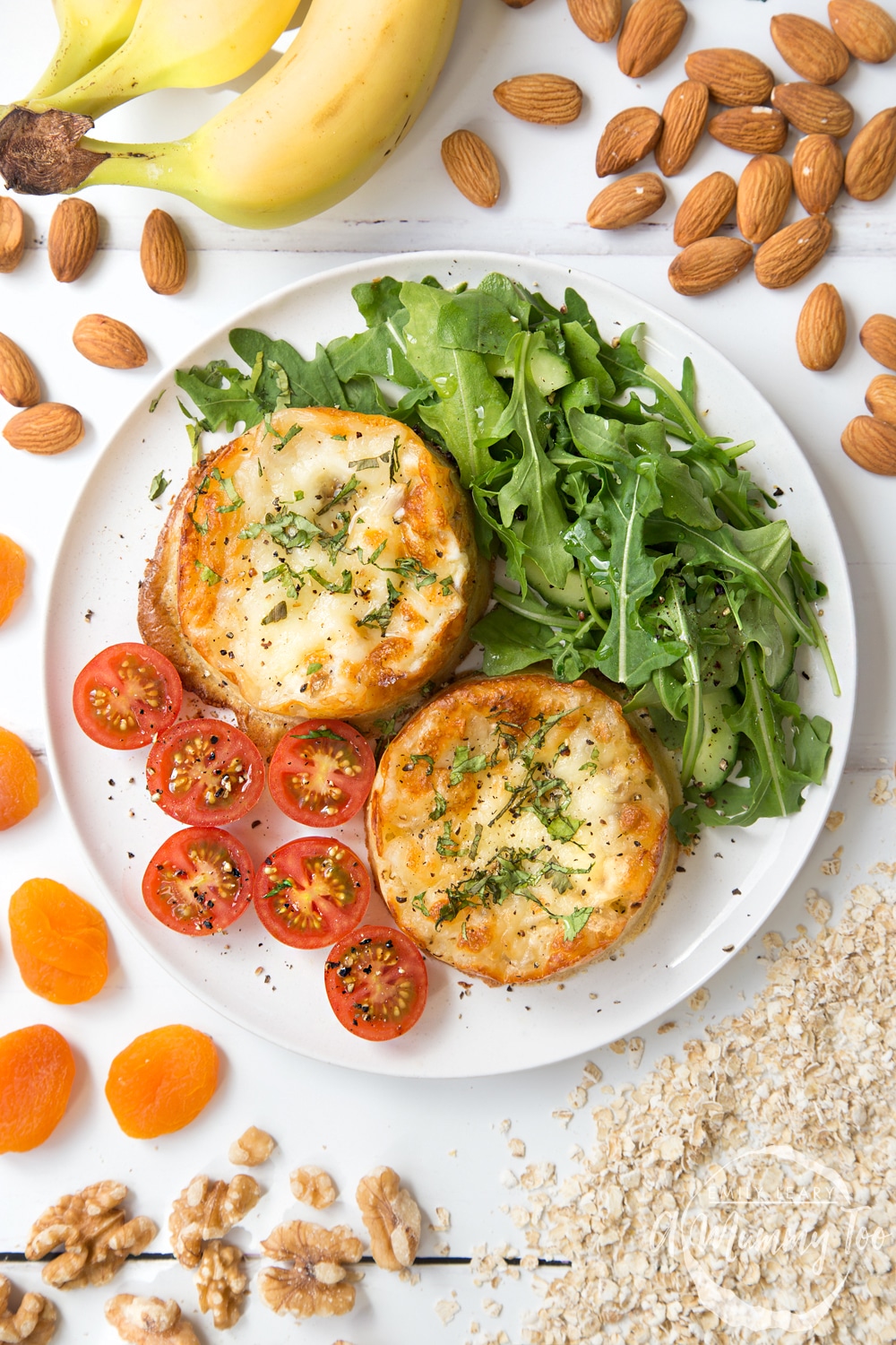 Overhead shot of a white plate with two Baked eggy crumpets. At the side of the plate there's some chopped cherry tomaotes and rocket salad.