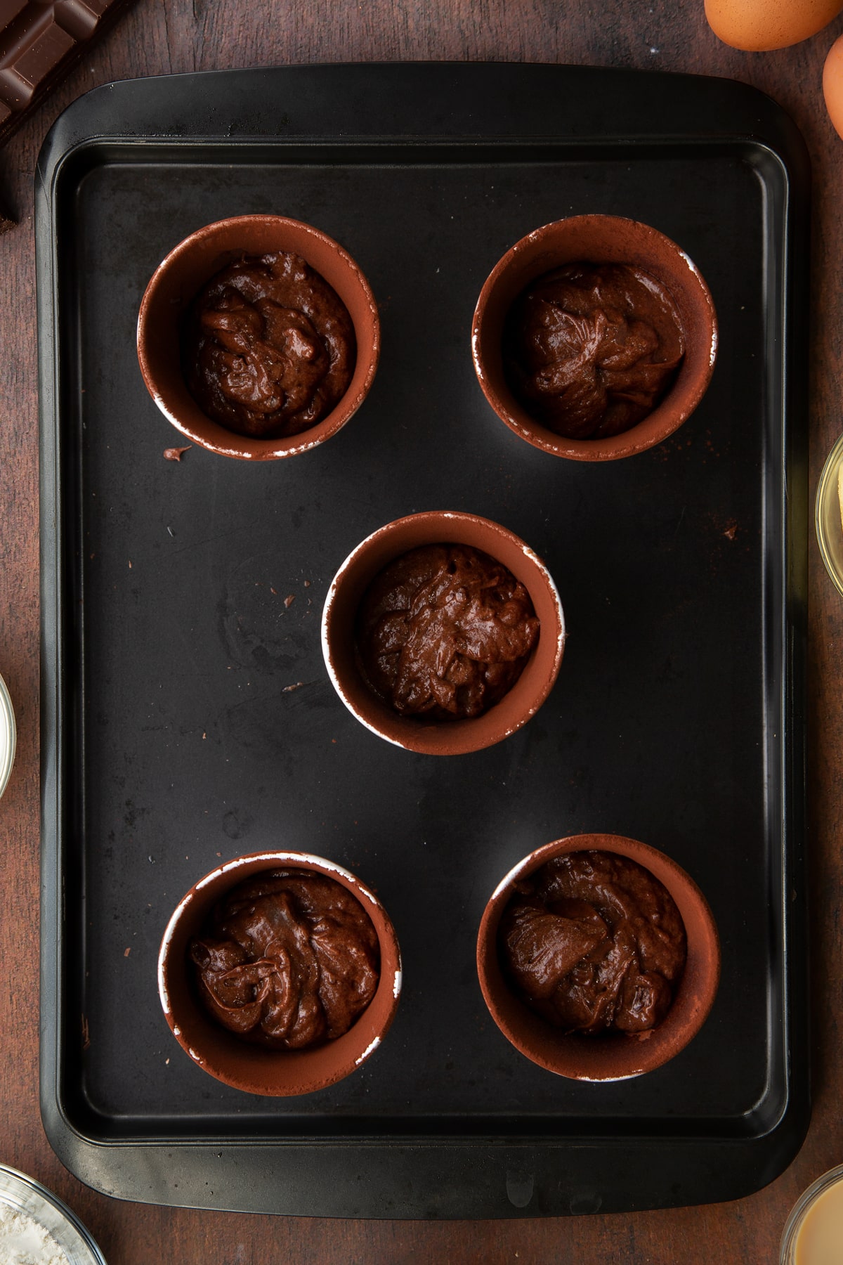 5 ceramic cake moulds filled with chocolate baileys cake mix on a baking tray.