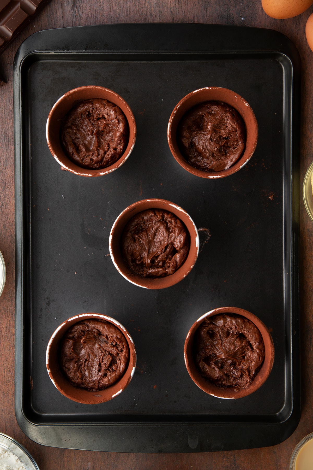 5 ceramic cake moulds filled with chocolate baileys cake mix on a baking tray.
