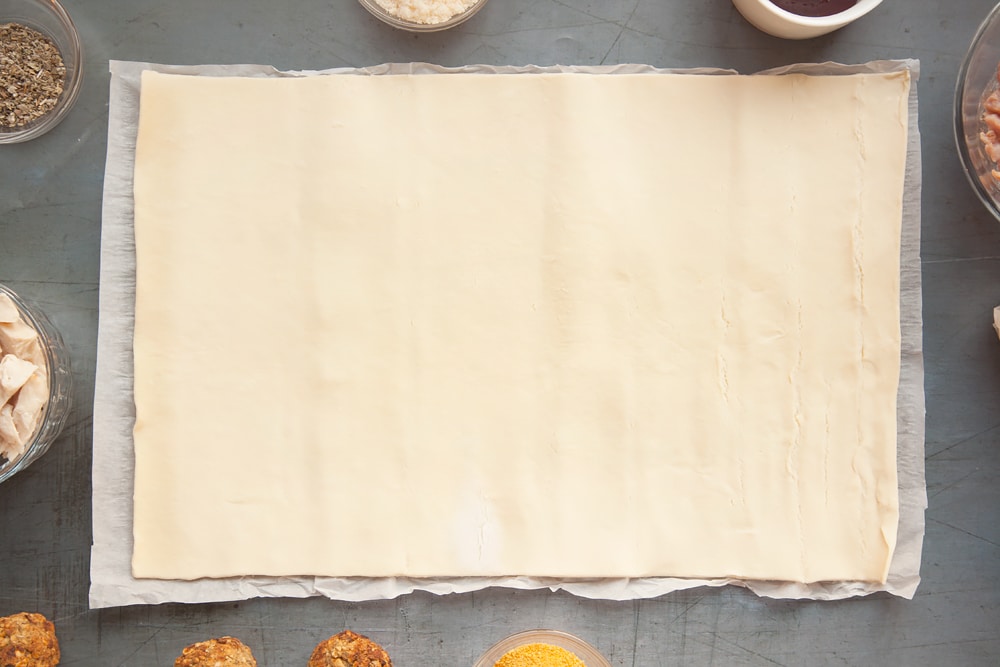 Unrolling a sheet of pasry onto a grey background. At the edge of the image there's additional bowls of ingredients.