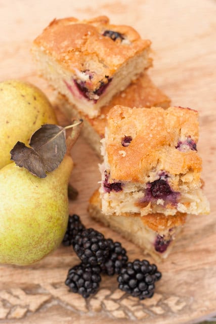 squares of Blackberry and Pear Traybake on a wooden board next to fresh pears and blackberries.