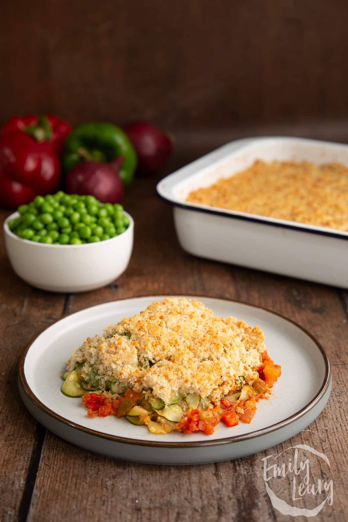 a piece of tomato and courgette gratin on a white plate with peas in a bowl in the background.