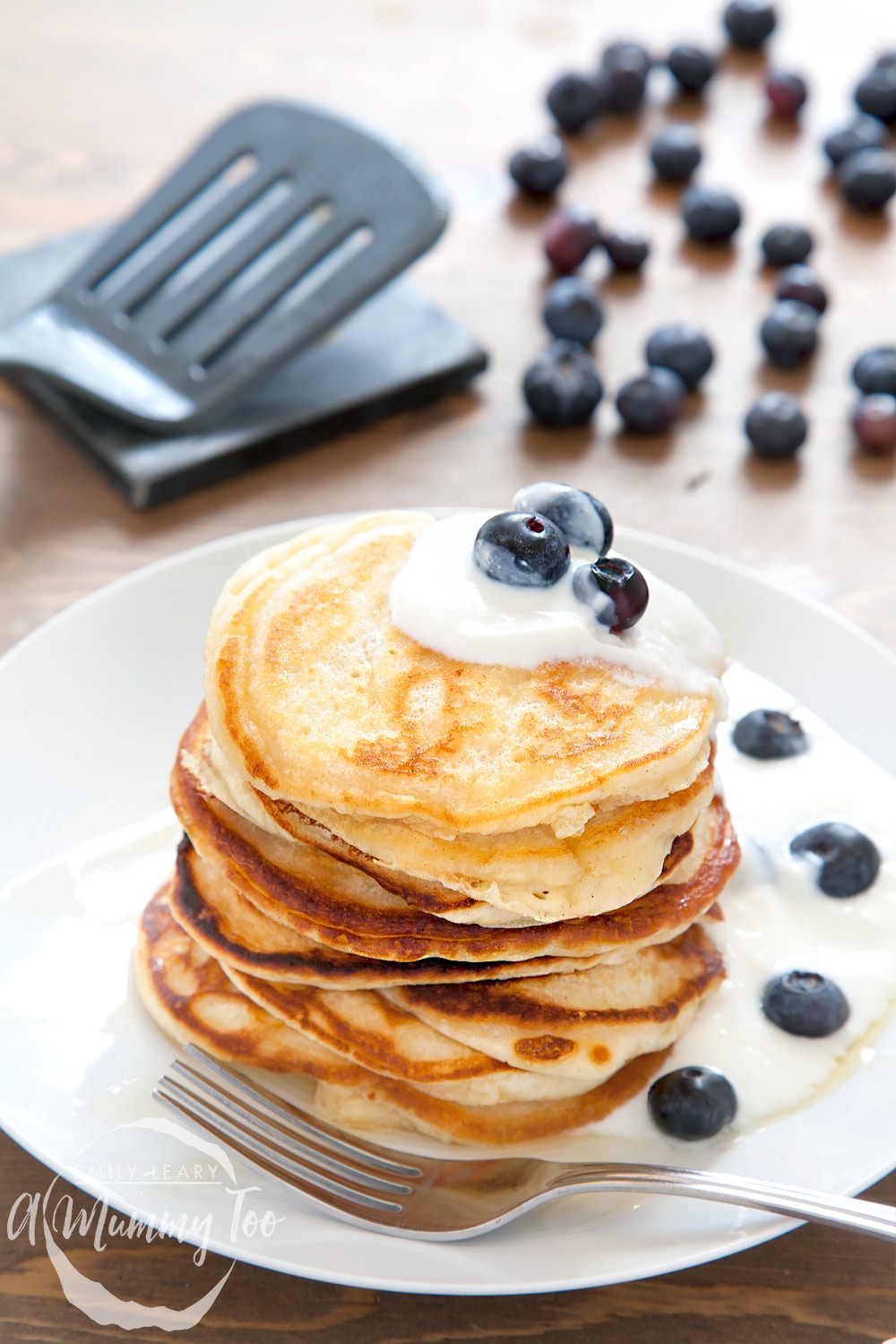 A stack of coconut yogurt pancakes, topped with coconut yogurt and blueberries with blueberries in the background.