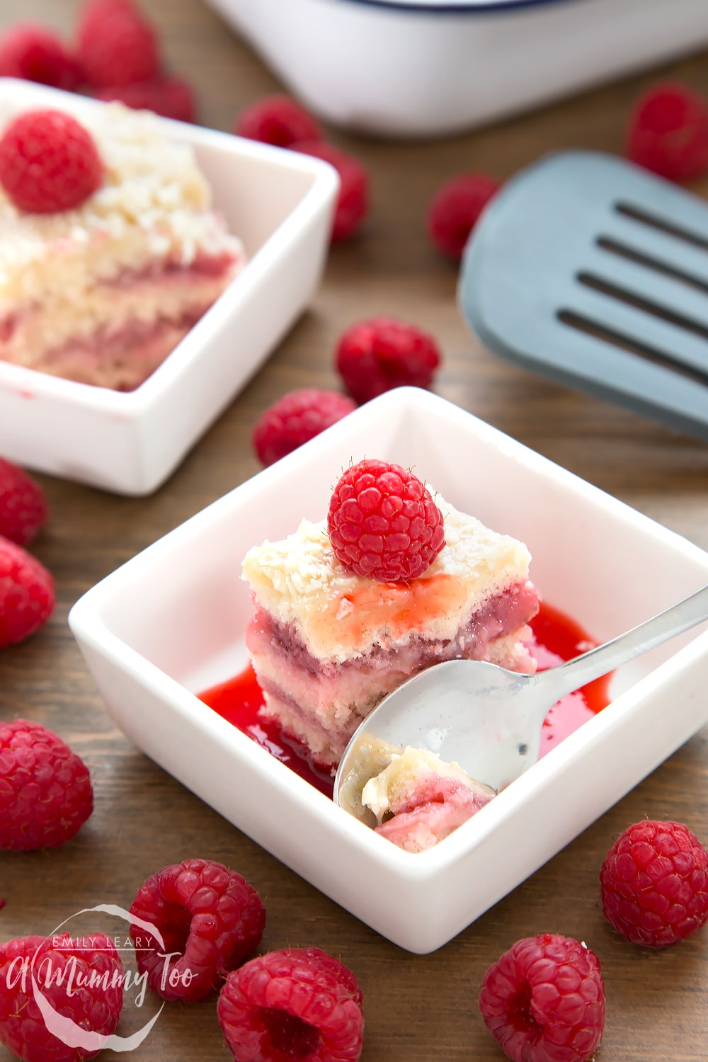 A piece of Strawberry and custard layer cake in a square white bowl, spoon on the side topped with a raspberry.