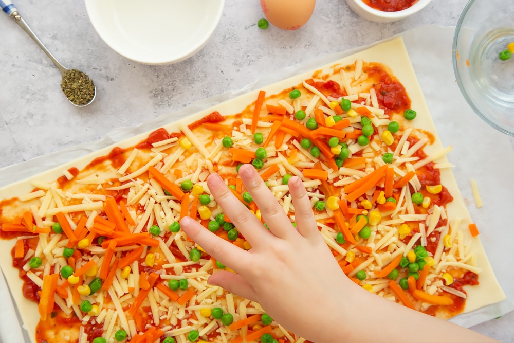 Sprinkling veggies on top of the pizza base, ready to create the veggie pizza roll-ups