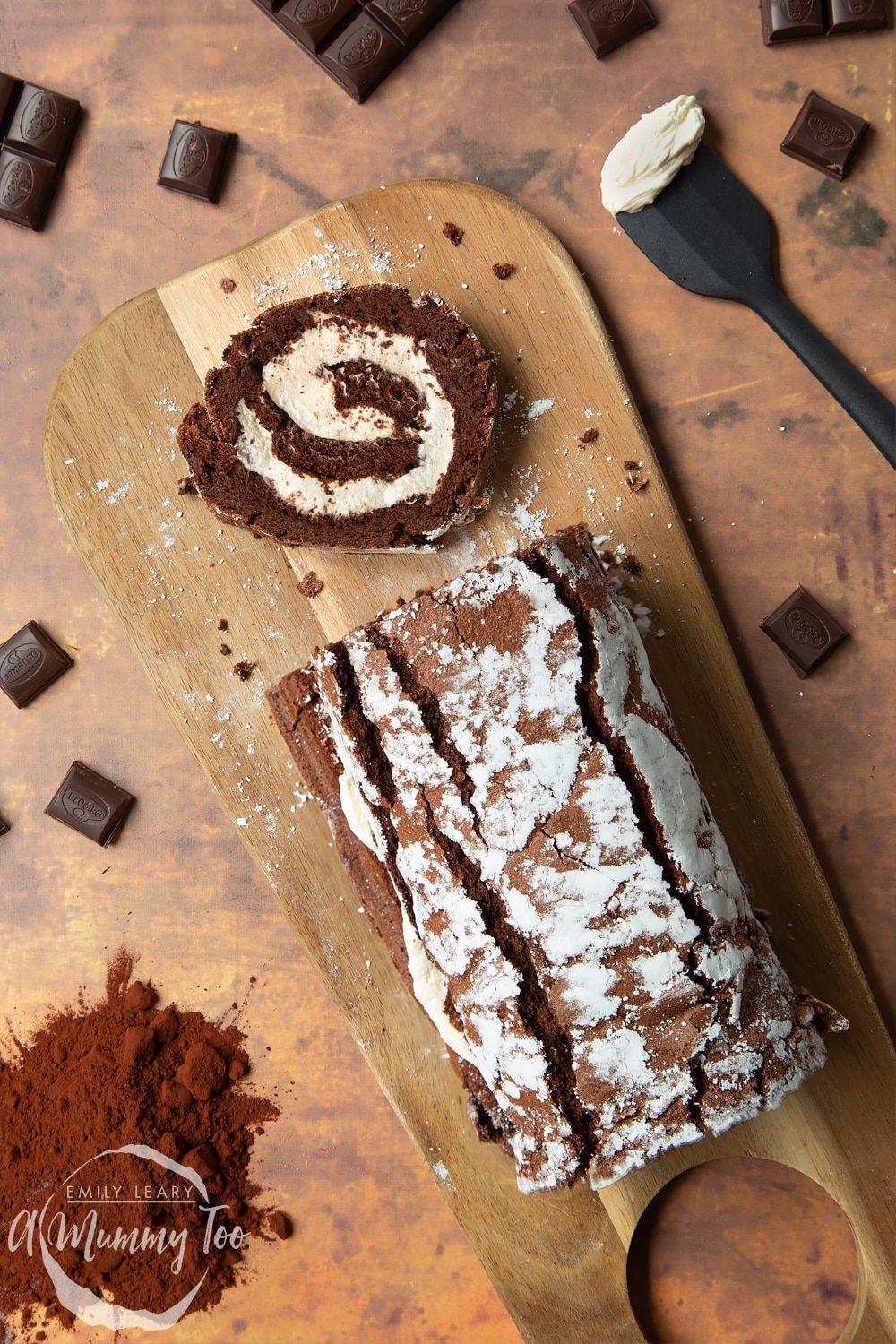 A chocolate roulard sits on a wooden chopping board. One slice of the chocolate roulard has been cut off. The surrounding areas is covered in dark chocolate squares.