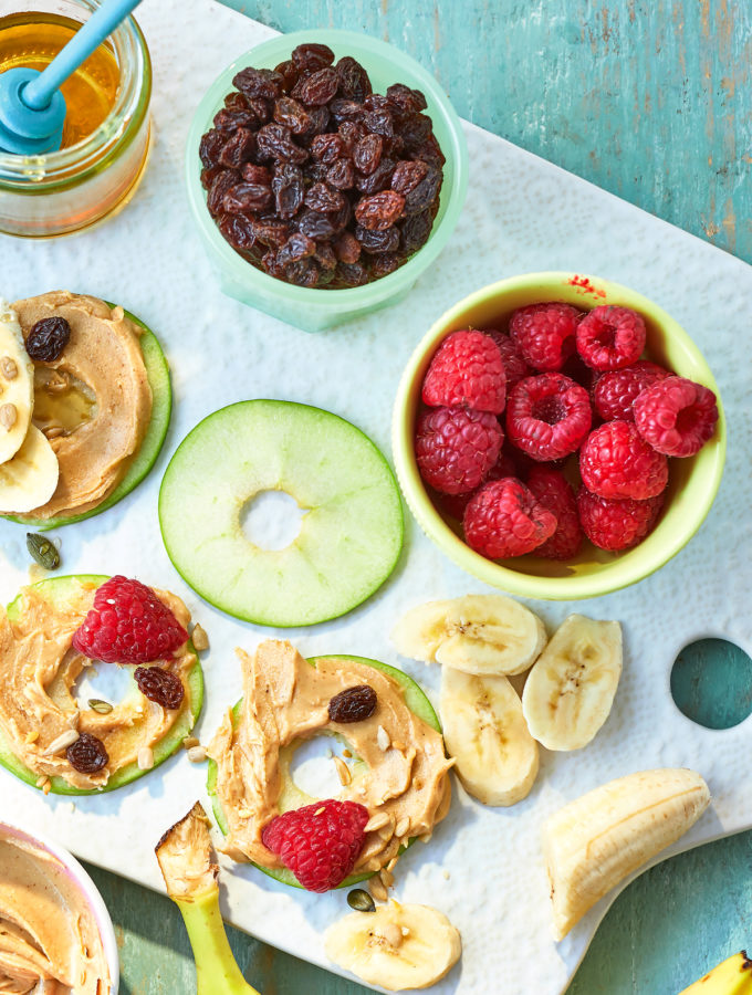 Overhead view of doughnut shaped apple slices and bowls of fruit. All but one of the apple slices are covered with peanut butter and more fruit.
