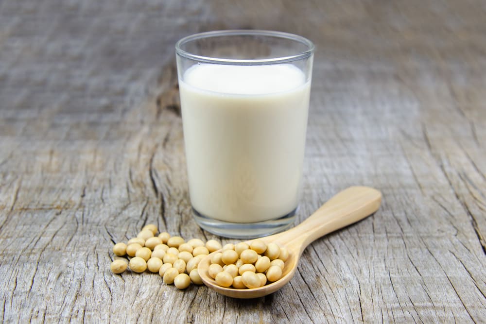 A glass of soy milk on a rustic wooden table. In front of the glass is some soy beans on a wooden spoon.