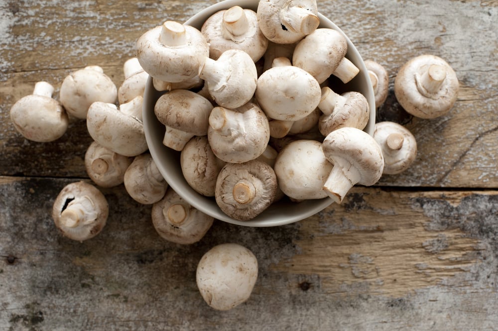 Overhead shot of mushrooms in a bowl. Not all of the mushrooms fit into the bowl and as a result some have spilled out onto the wooden table.