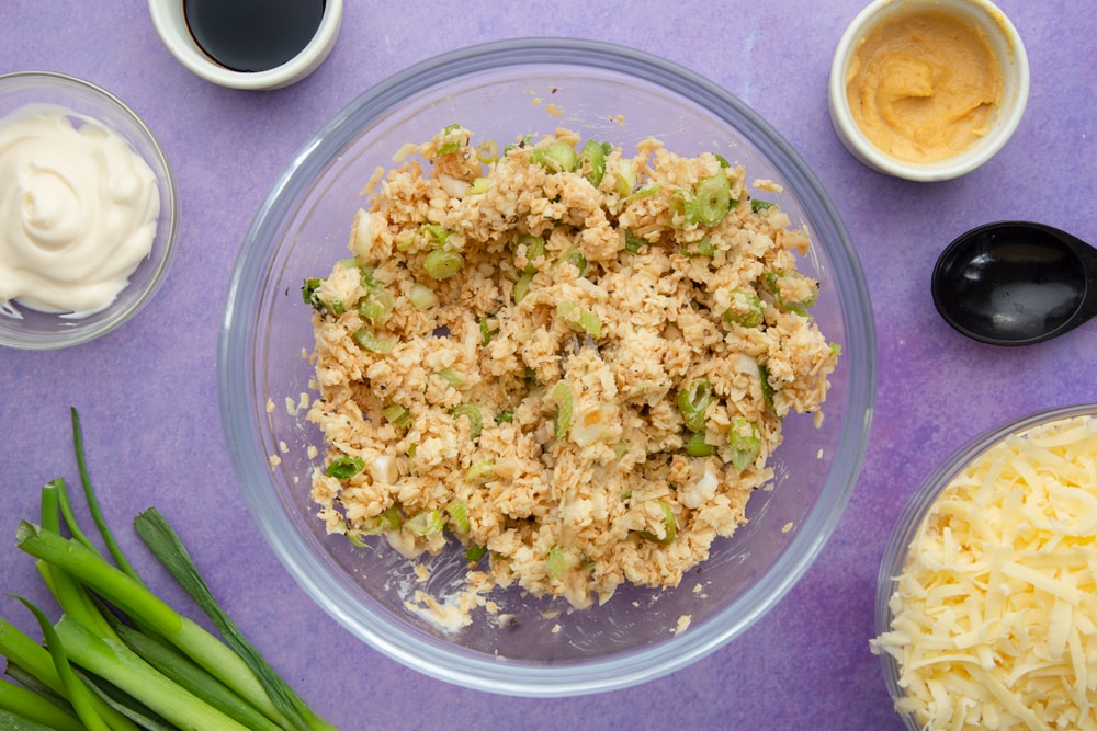 Grated mozzarella, cheddar, spring onions, mayonnaise, mustard and soy sauce mixed together in a mixing bowl. Ingredients to make cheese and onion jacket potatoes surround the bowl.