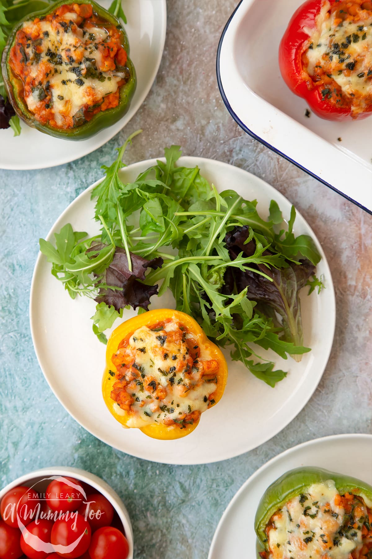 A yellow pepper stuffed with turkey mince, vegetables and baked beans with melted cheese on top on a white plate. More baked bean stuffed peppers are shown to the edge of frame.