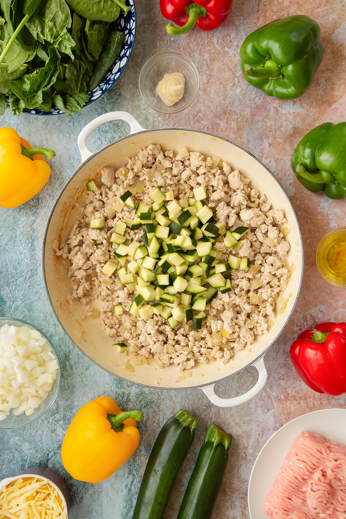 Lightly fried onions, garlic and turkey mince in a large shallow pan with chopped courgette on top. Ingredients to make baked bean stuffed peppers surround the pan.