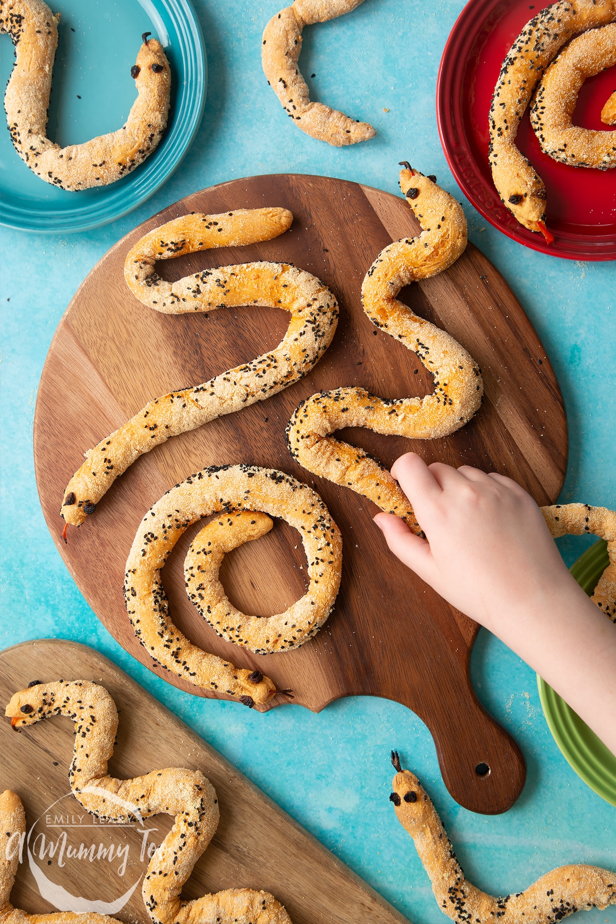 Bread snakes on a wooden board. More bread snakes are shown on coloured plates at the edges of the photo. A hand reaches to take a snake.