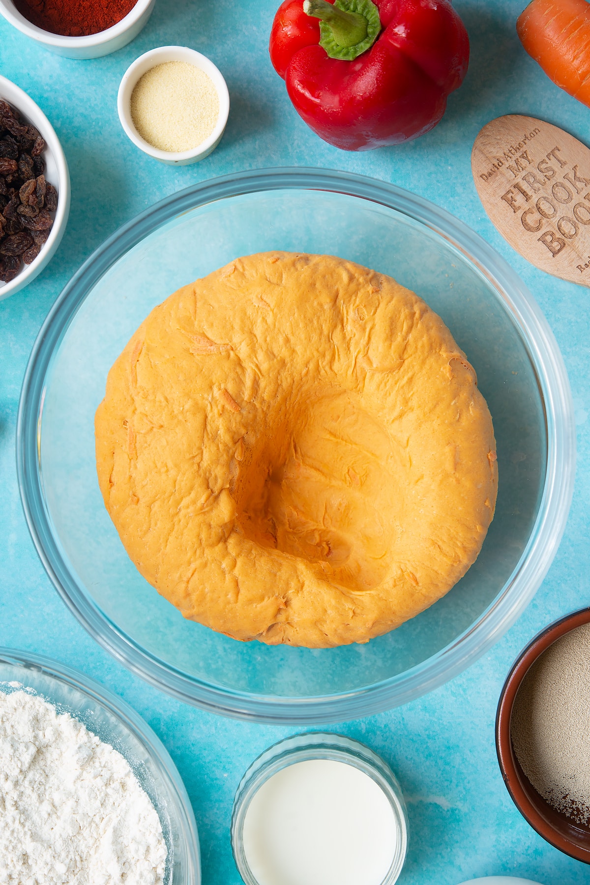 Punched back paprika and carrot bread dough in a bowl. Ingredients to make bread snakes surround the bowl.