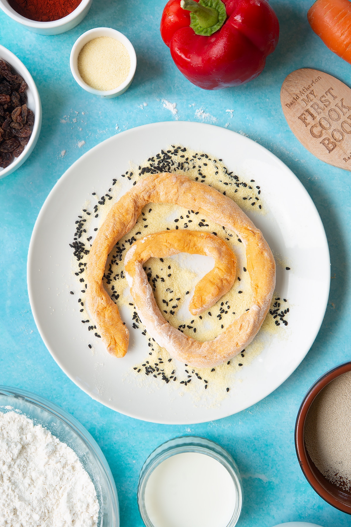 Paprika and carrot bread dough rolled into a long sausage on a plate with semolina and nigella seeds. Ingredients to make bread snakes surround the plate.