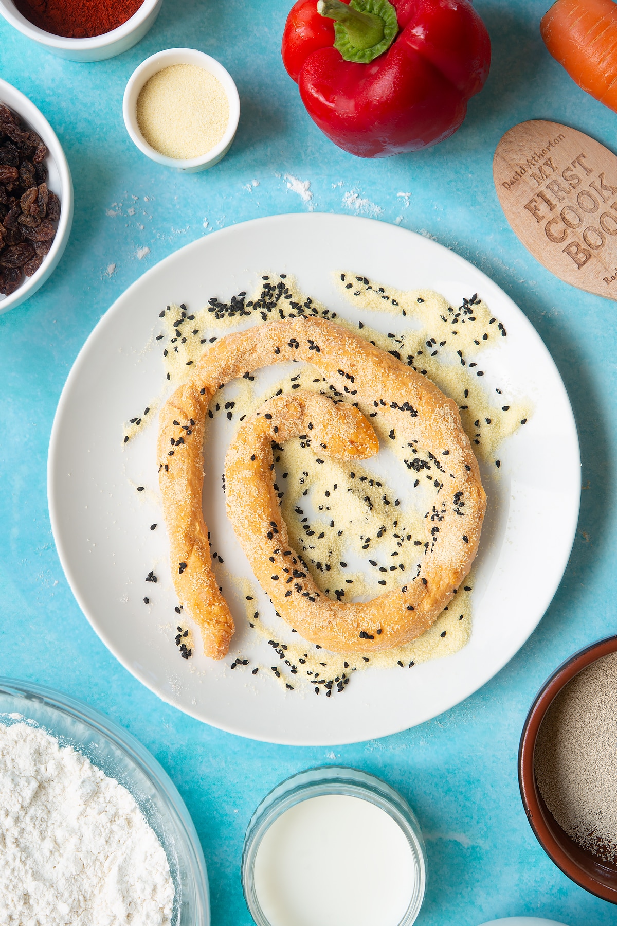 Paprika and carrot bread dough rolled into a long sausage on a plate, rolled in semolina and nigella seeds. Ingredients to make bread snakes surround the plate.