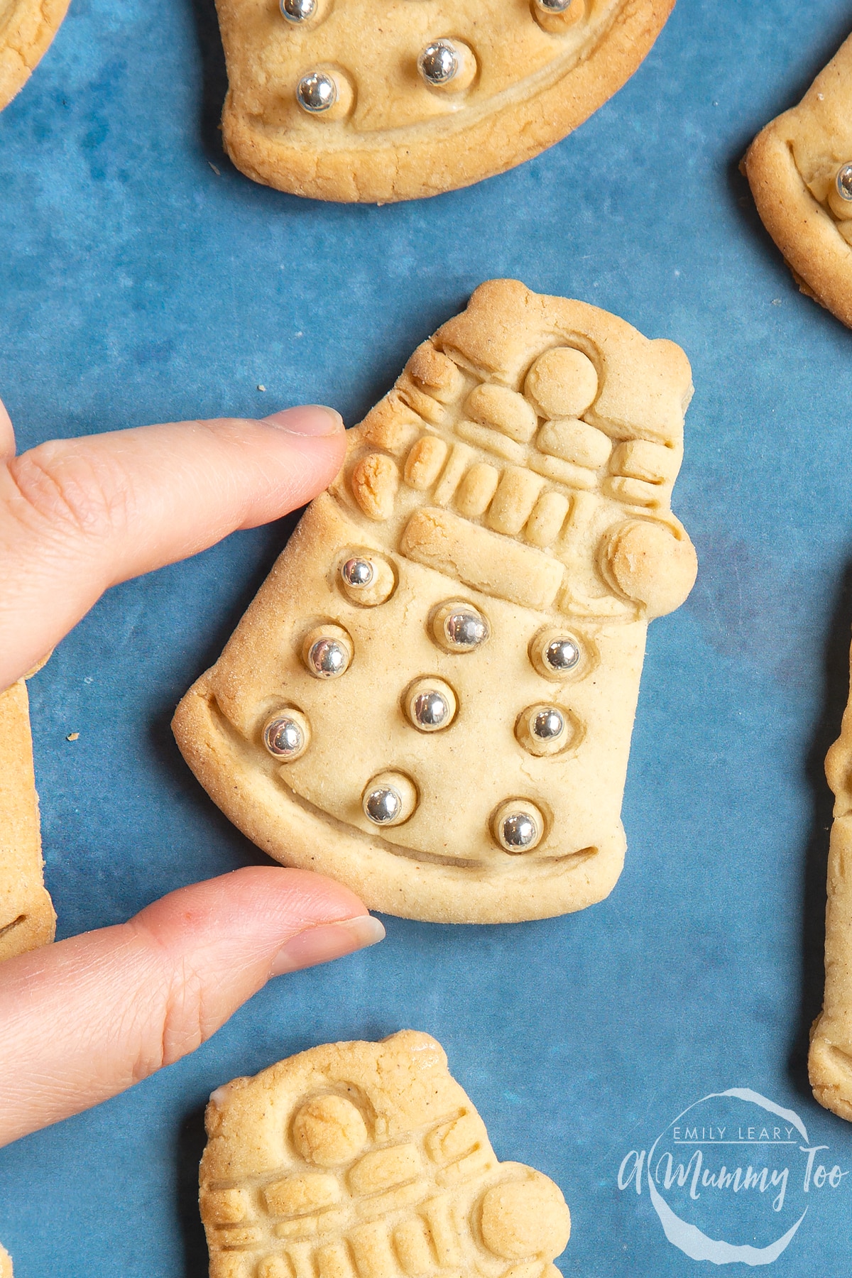 Close up of a Dalek cookies on a blue background. The cookie is decorated with edible silver candy balls. A hand reaches to take it.