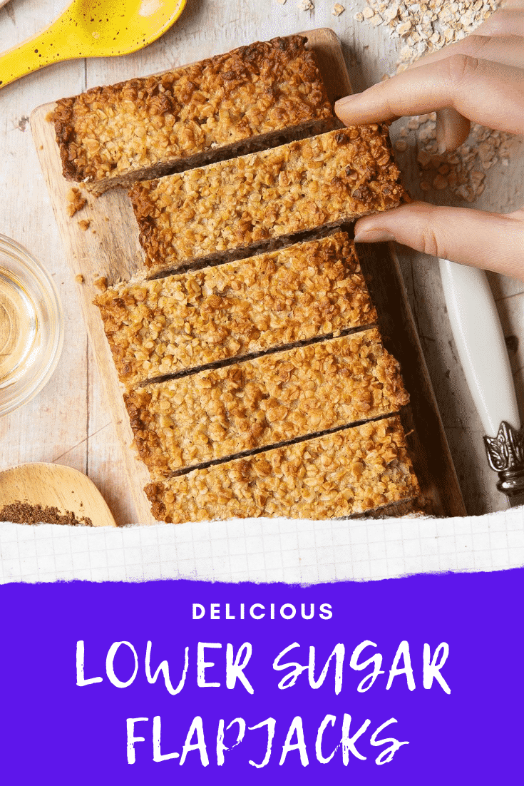 Overhead shot of a hand holding a lower sugar flapjack served on a wooden plate with the delicious low sugar graphic at the bottom.