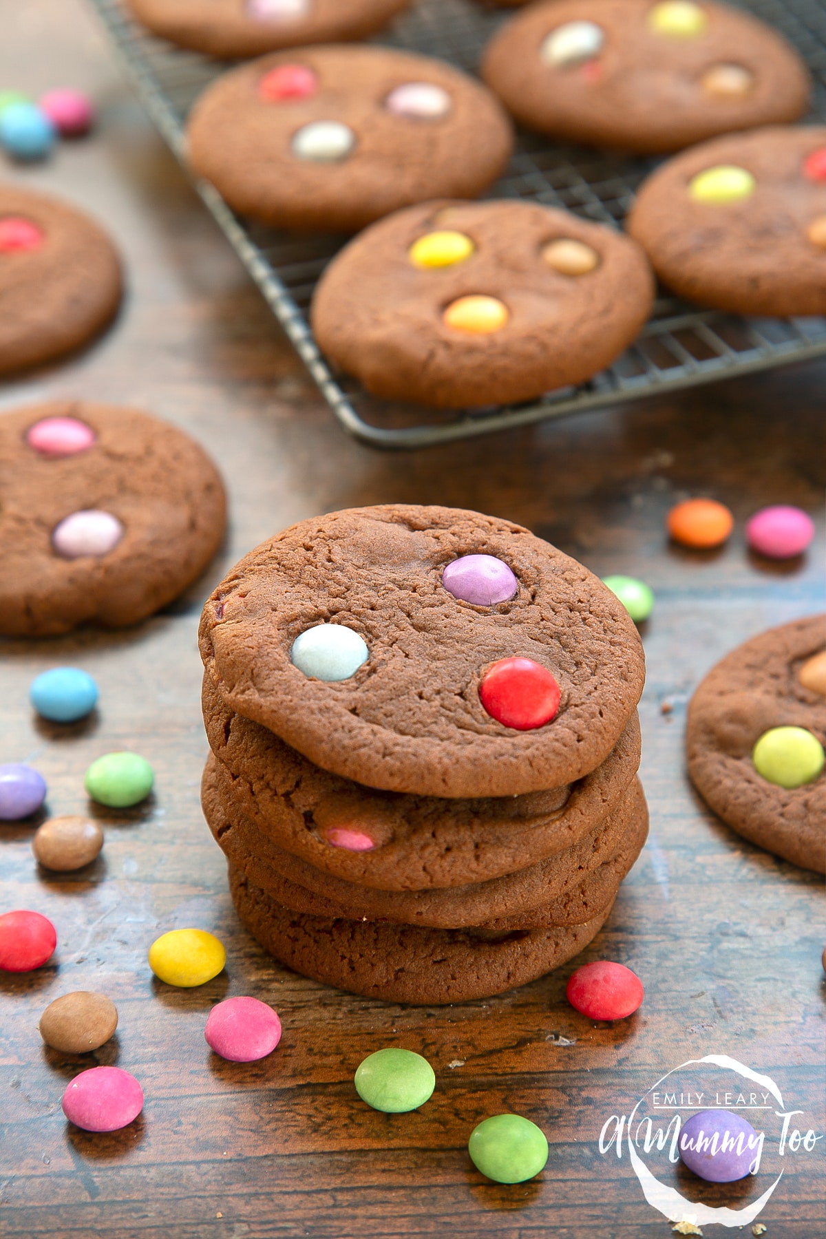 Smartie cookies stacked up on a dark wooden surface. The cookies are surrounded by scattered Smarties.