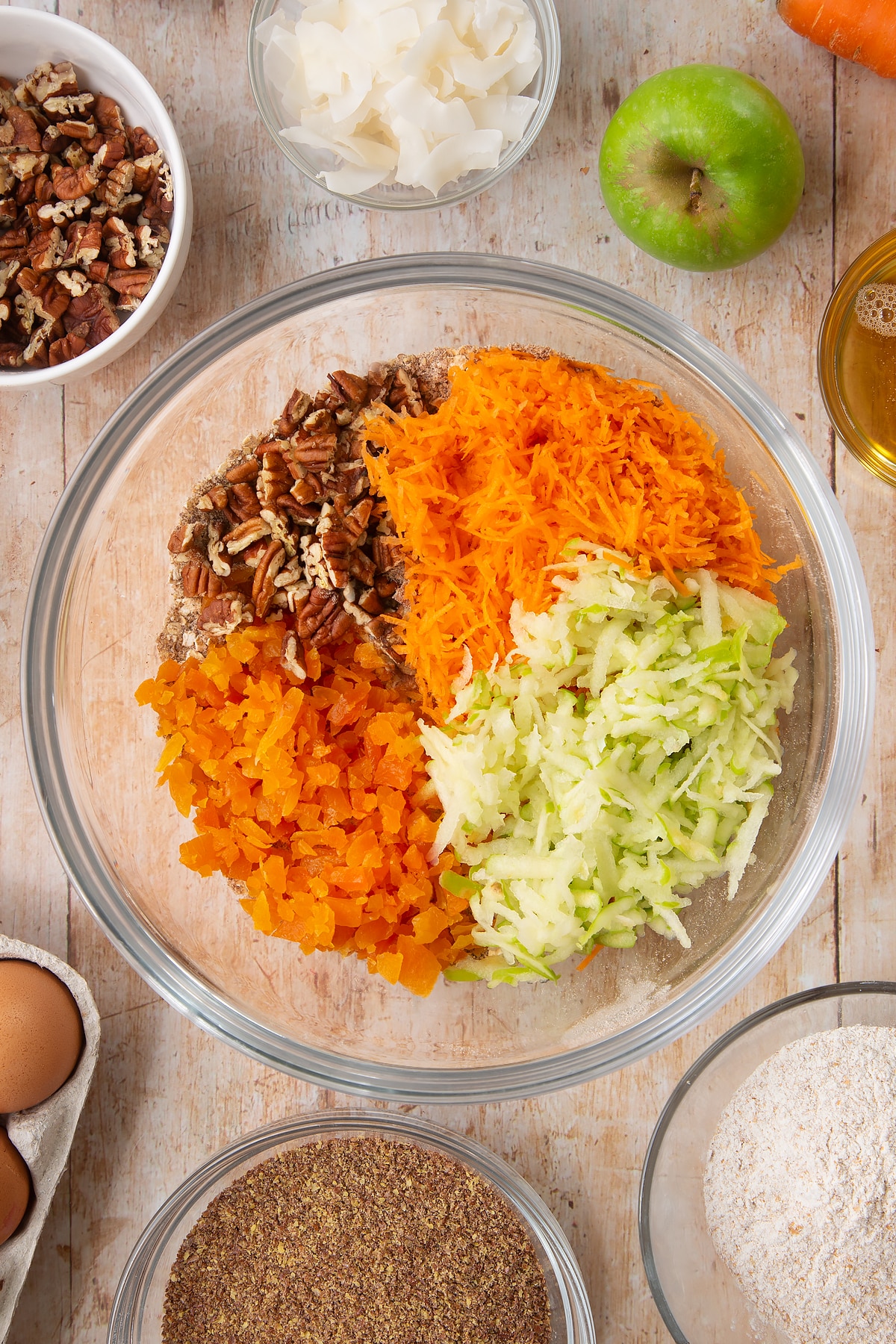 Overhead shot of dry ingredients mix, dried apricots, nuts, apple and carrot in a large clear bowl