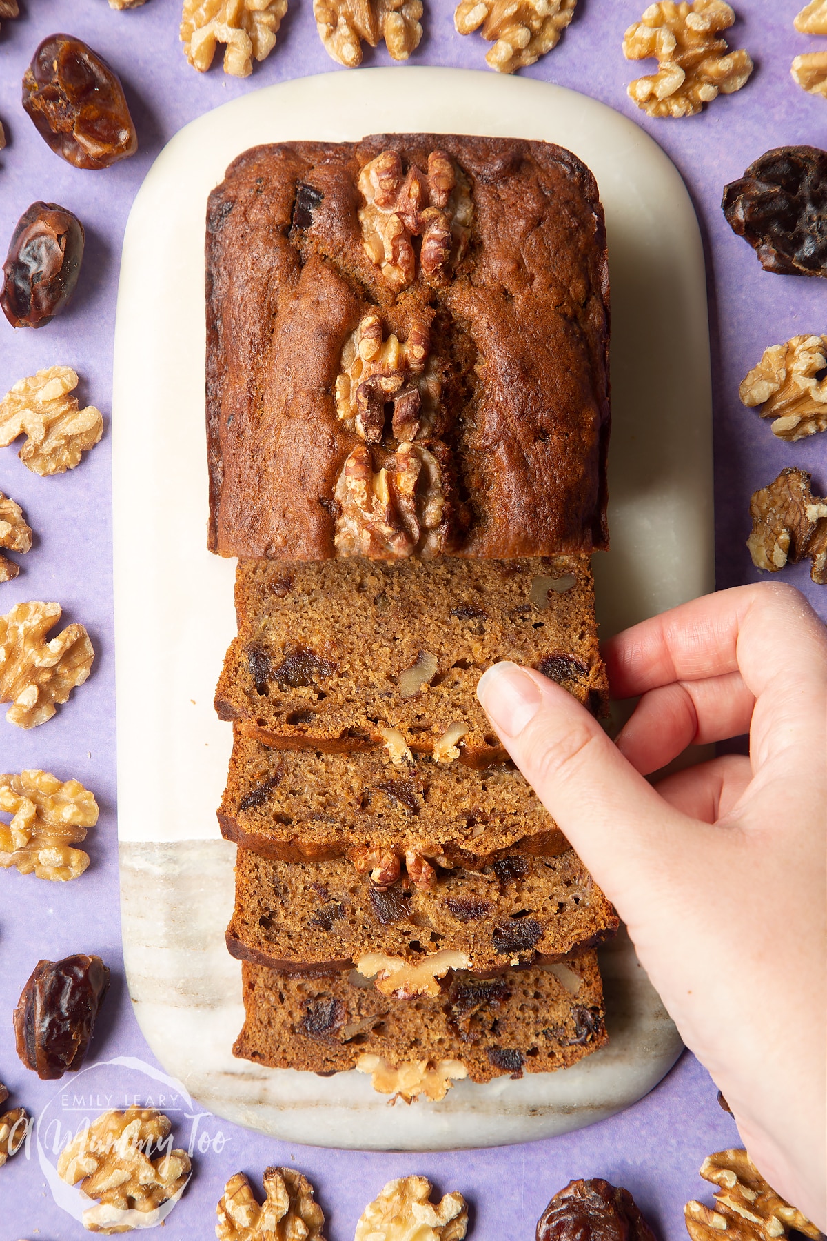 Overhead shot of a hand holding a Vegan date and walnut cake served on a marble plate with a mummy too logo in the lower-left corner