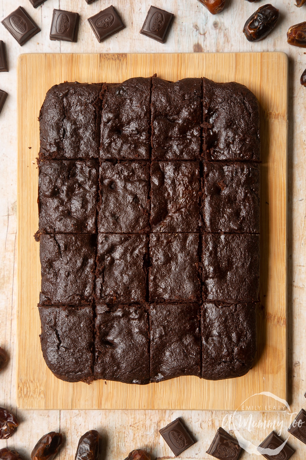 16 whole grain brownies made with dates and dark chocolate arranged on a pale wooden board.