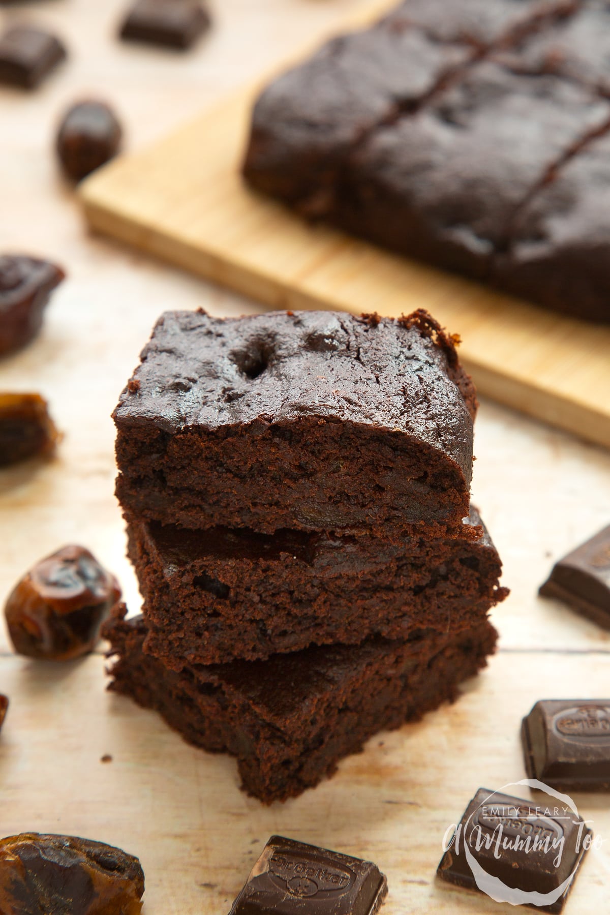 Whole grain brownies made with dates and dark chocolate, stacked up on pale wood. Squares of dark chocolate and dates surround the brownies.
