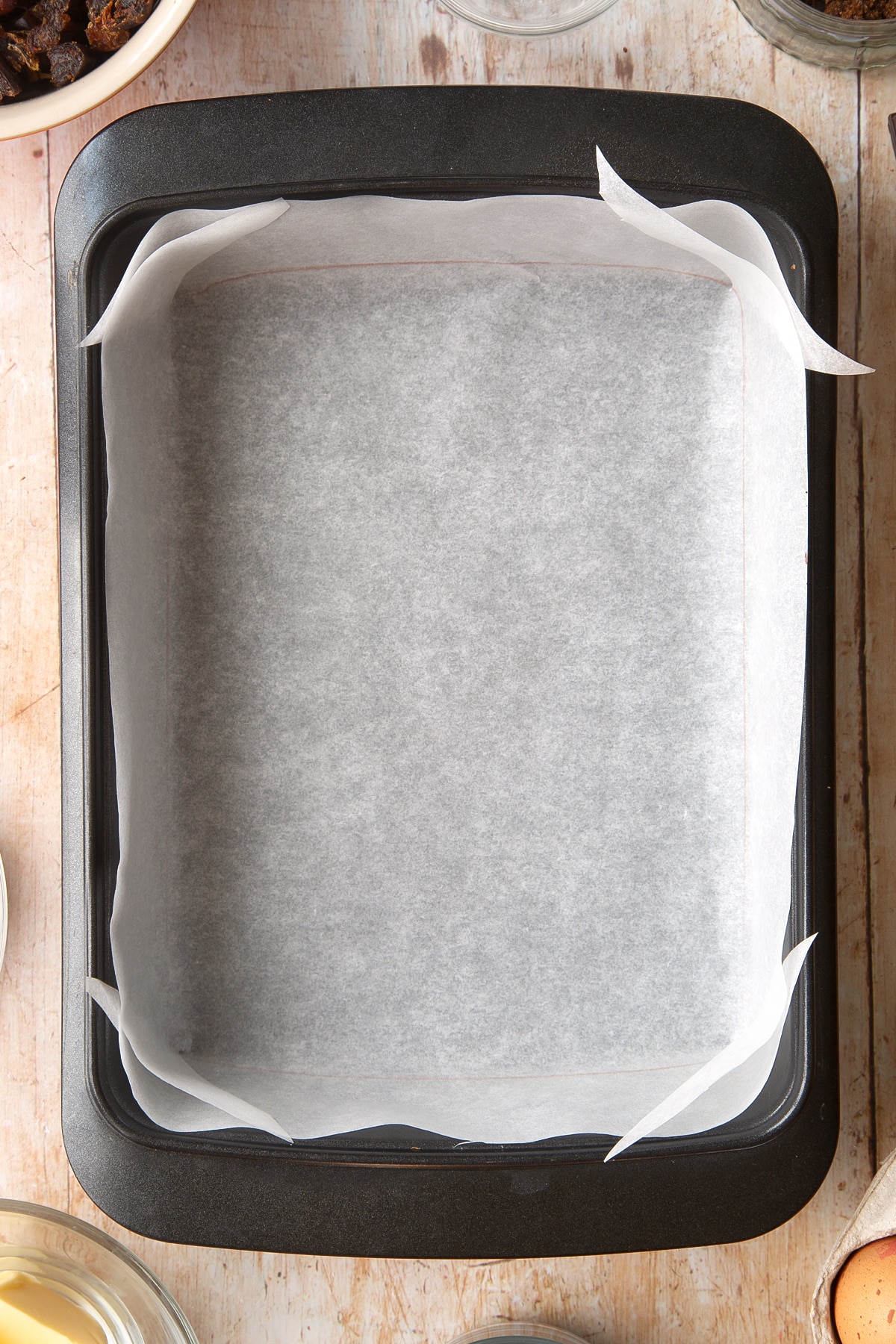 A rectangular tin lined with baking paper. Ingredients to make whole grain brownies surround the tin.