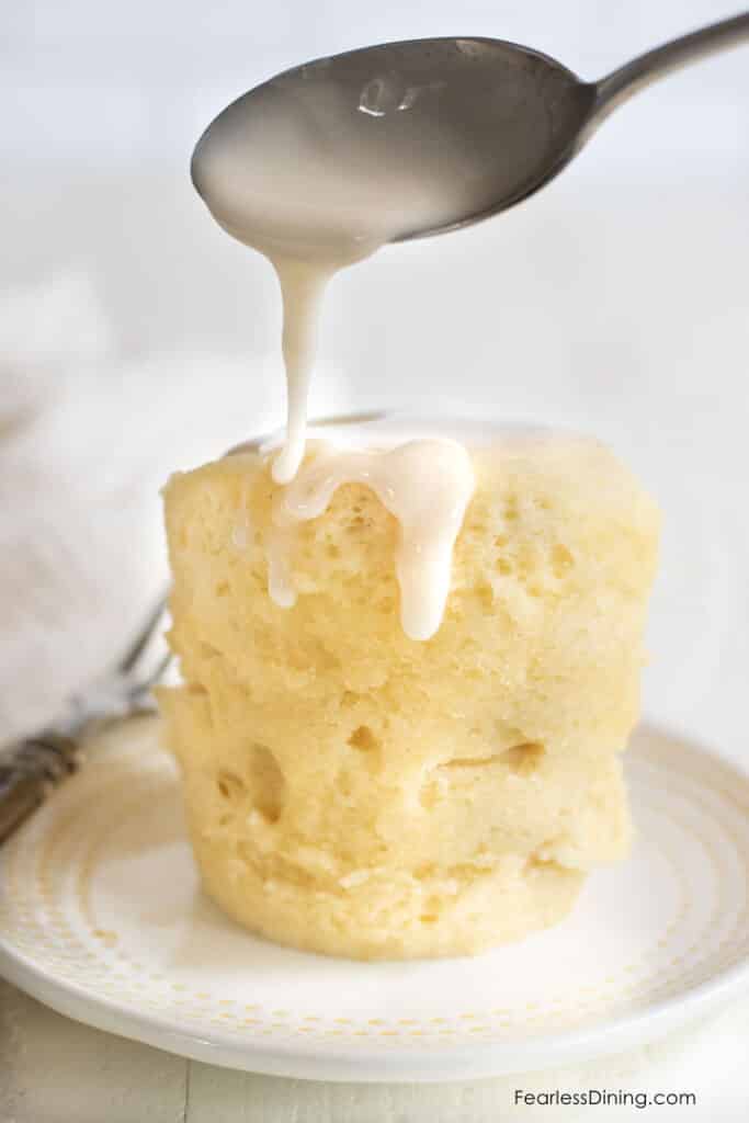 Gluten free lemon mug cake served onto a small plate. A spoon drizzles lemon icing over the cake.