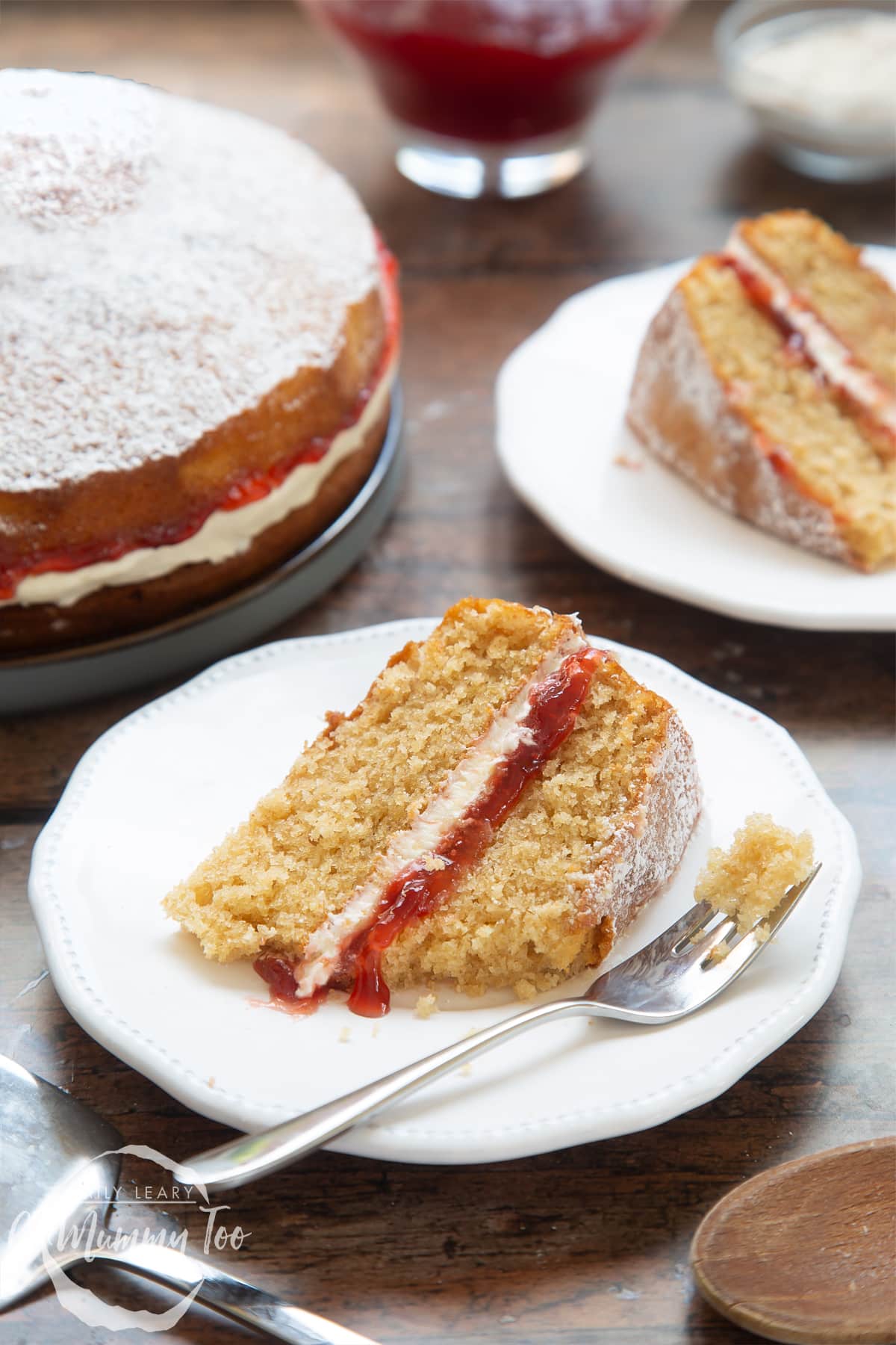 A slice of golden syrup sponge cake on a white decorate plate with a cake fork on the side. In the background there's an additional slice of cake as well as the cake as a whole.