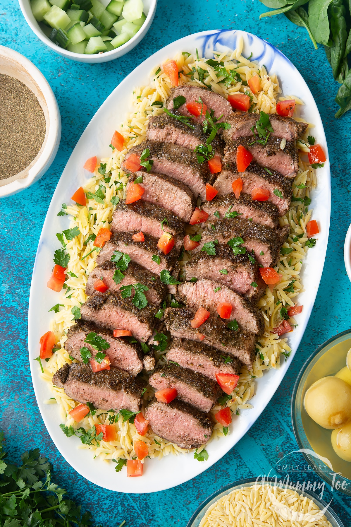 Slices of herby lamb arranged on a bed of orzo on an oval platter. The dish has been scattered with parsley and chopped tomatoes.