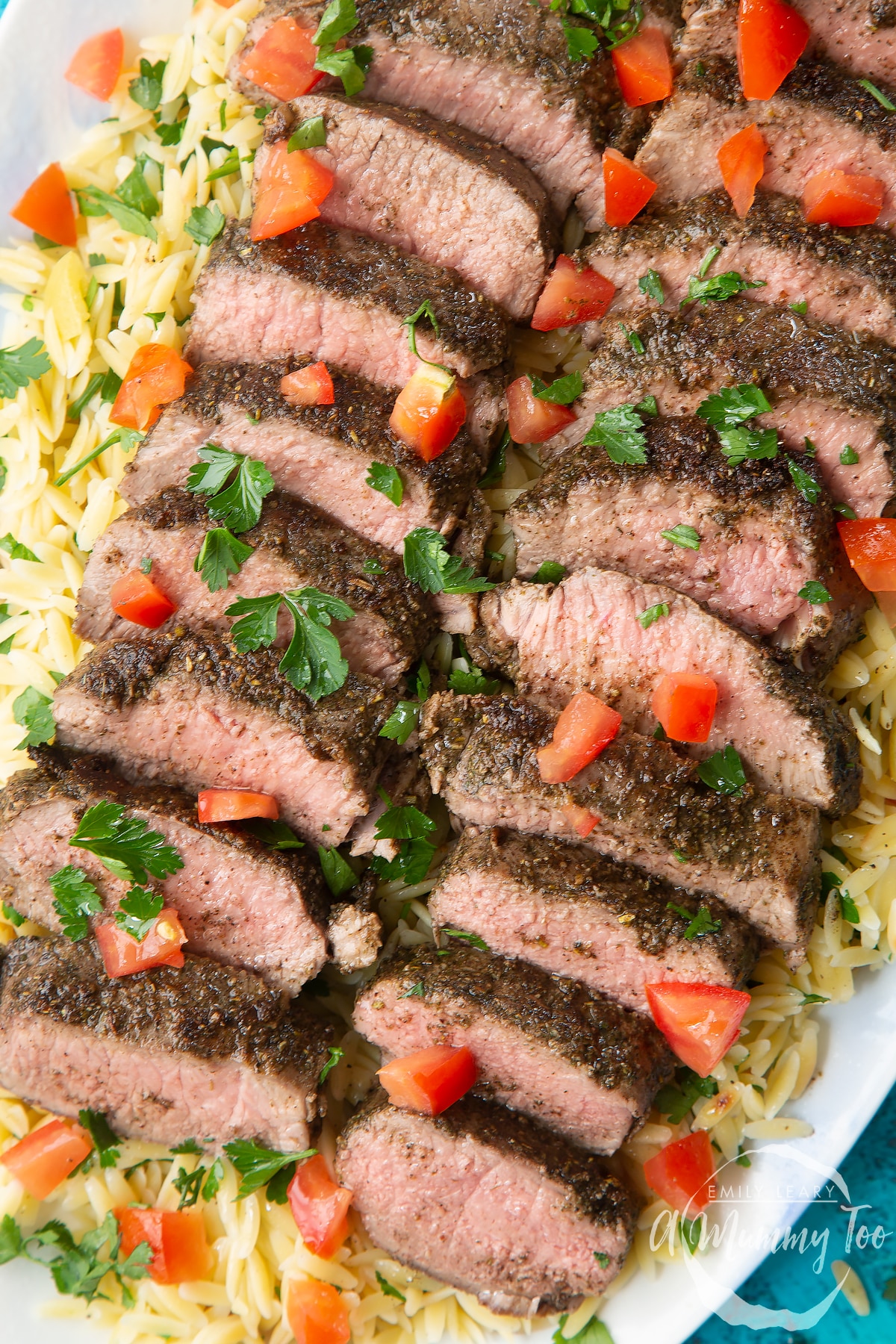 Close up of slices of herby lamb arranged on a bed of orzo on an oval platter. The dish has been scattered with parsley and chopped tomatoes.