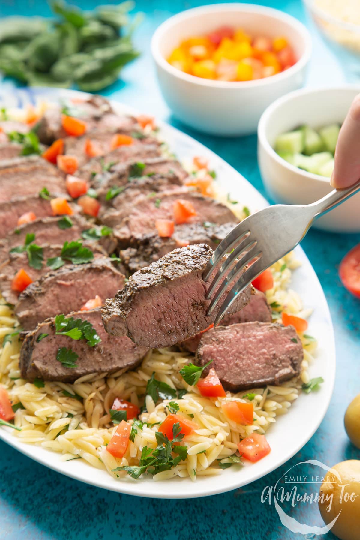 Slices of herby lamb arranged on a bed of orzo on an oval platter. The dish has been scattered with parsley and chopped tomatoes. A fork lifts a piece of lamb.