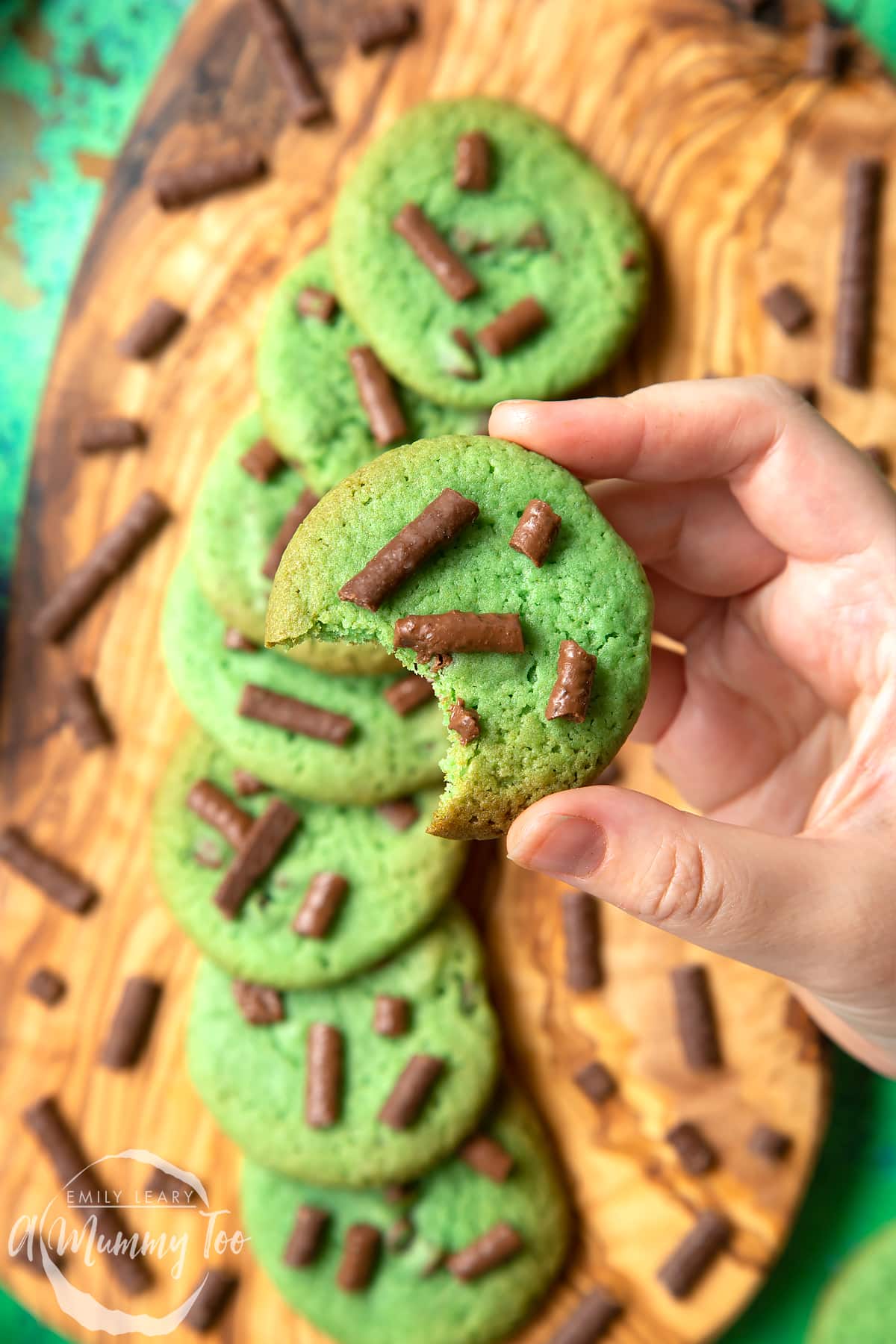 Hand holding a green cookie topped with Matchmaker pieces. It has a bite out of it. More Matchmaker cookies are arranged on a wooden board below.