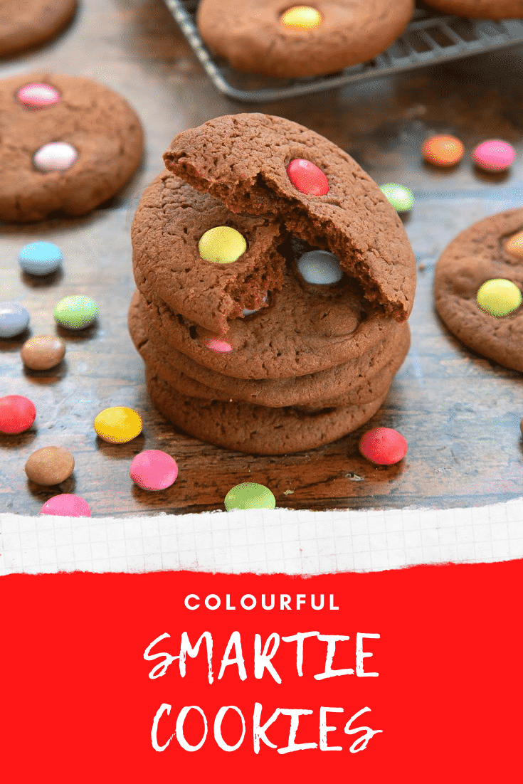 Smartie cookies stacked up on a dark wooden surface. The top cookie has been broken in two. Caption reads: colourful Smartie cookies