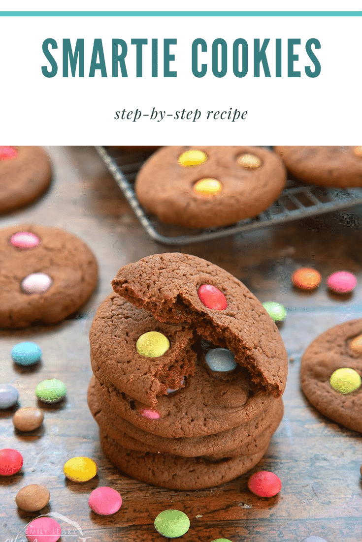 Smartie cookies stacked up on a dark wooden surface. The top cookie is broken in two. Caption reads: Smartie cookies step-by-step recipe
