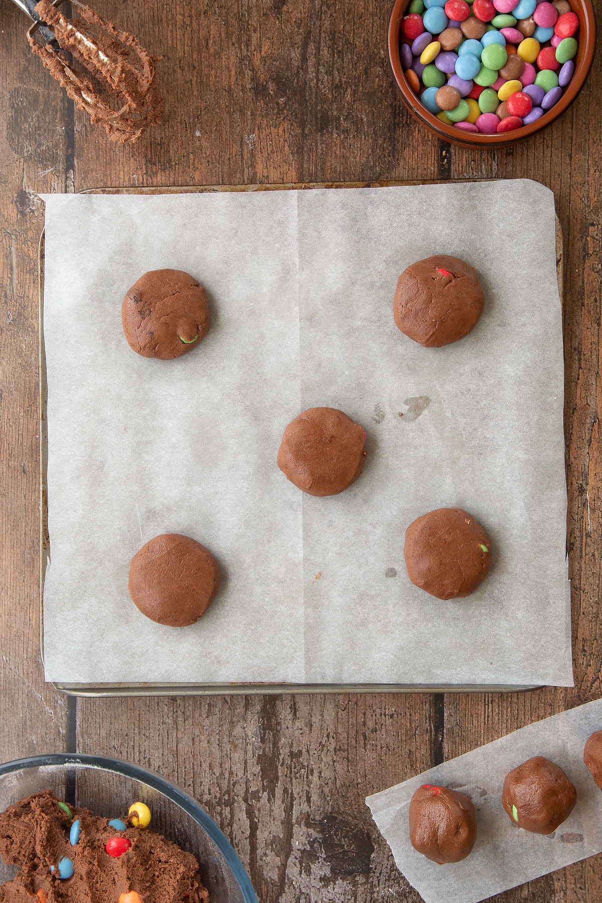 Smartie cookies on a baking tray lined with baking paper, ready to top with more Smarties. Ingredients to make the Smartie cookie recipe surround the tray.