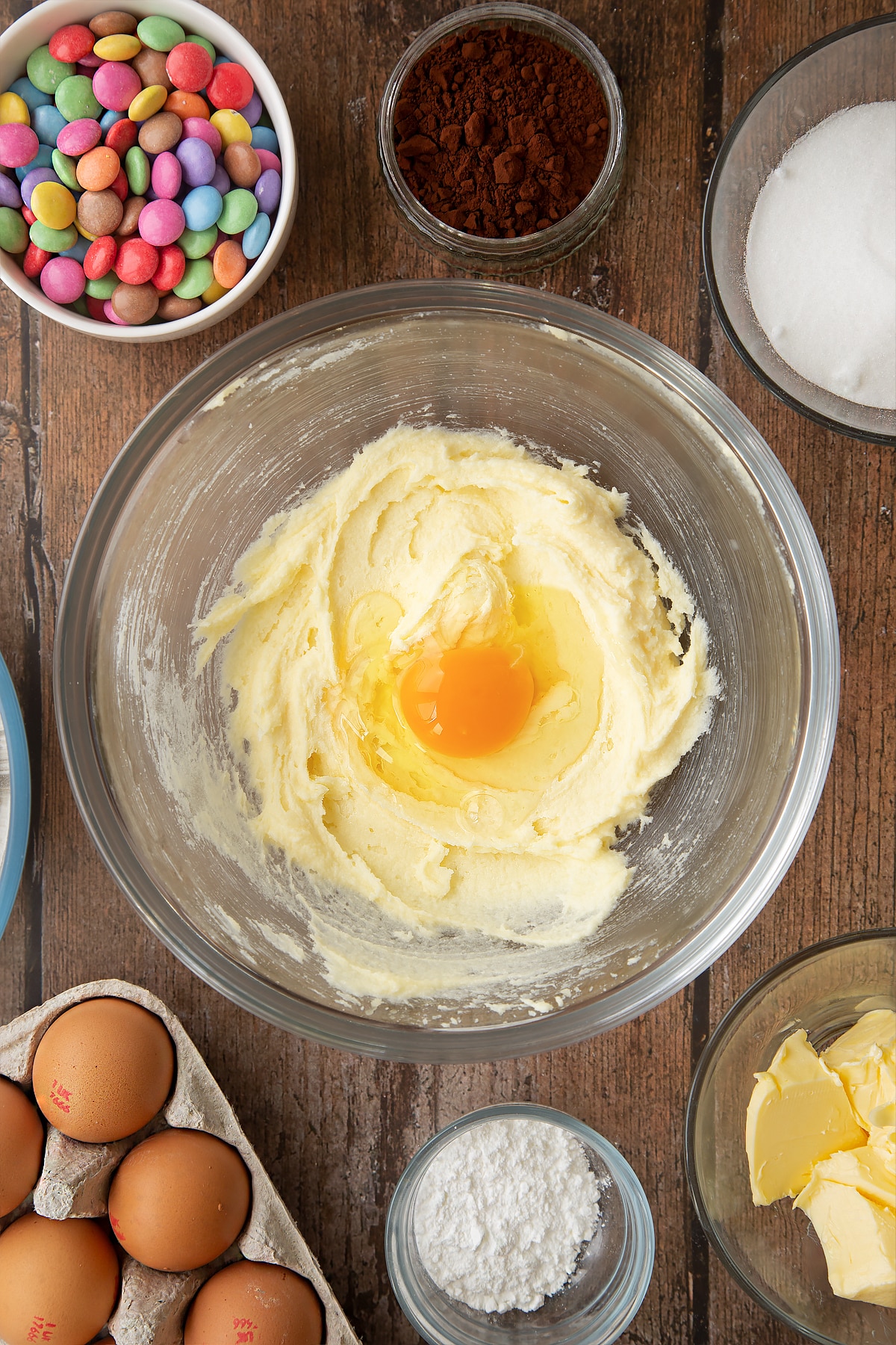 Beaten sugar and butter with an egg on top in a glass mixing bowl. Ingredients to make the Smartie cookie recipe surround the bowl.