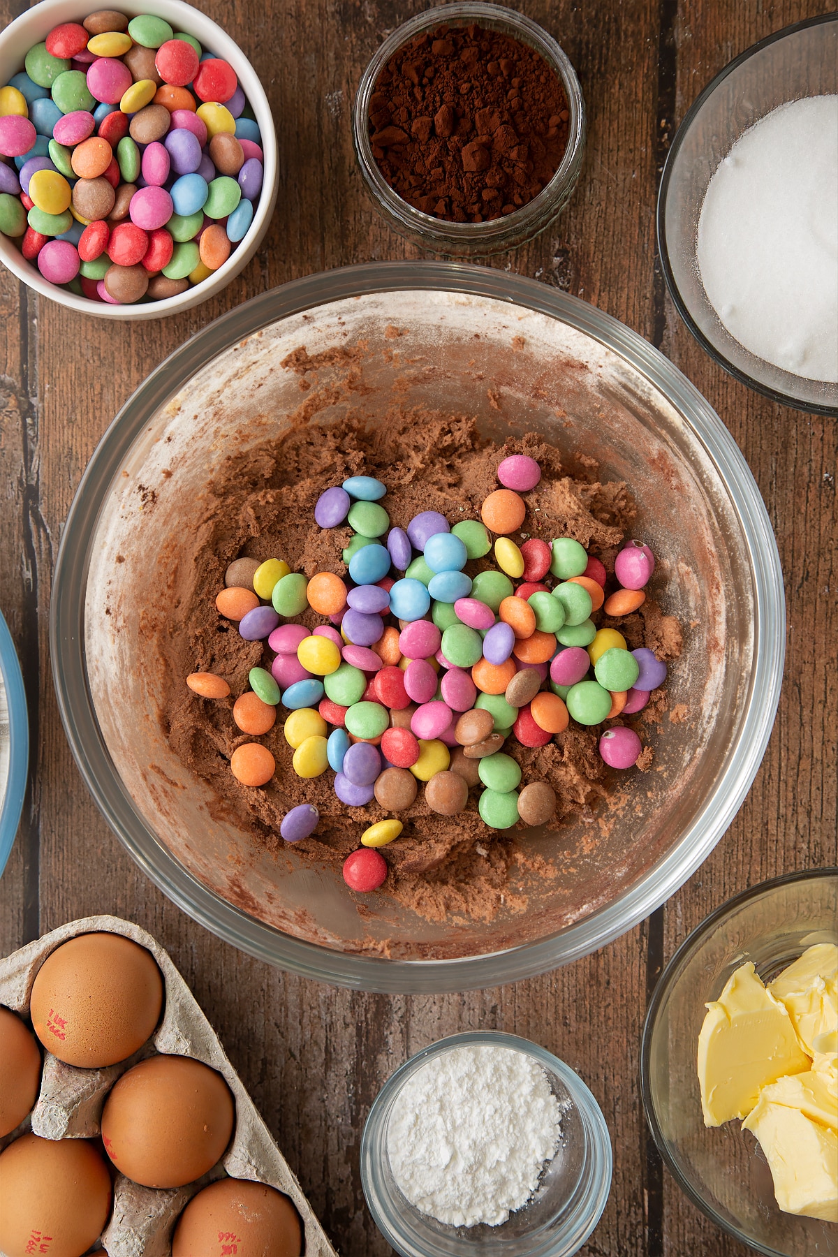 Chocolate cookie dough in a glass mixing bowl, topped with Smarties. Ingredients to make the Smartie cookie recipe surround the bowl.