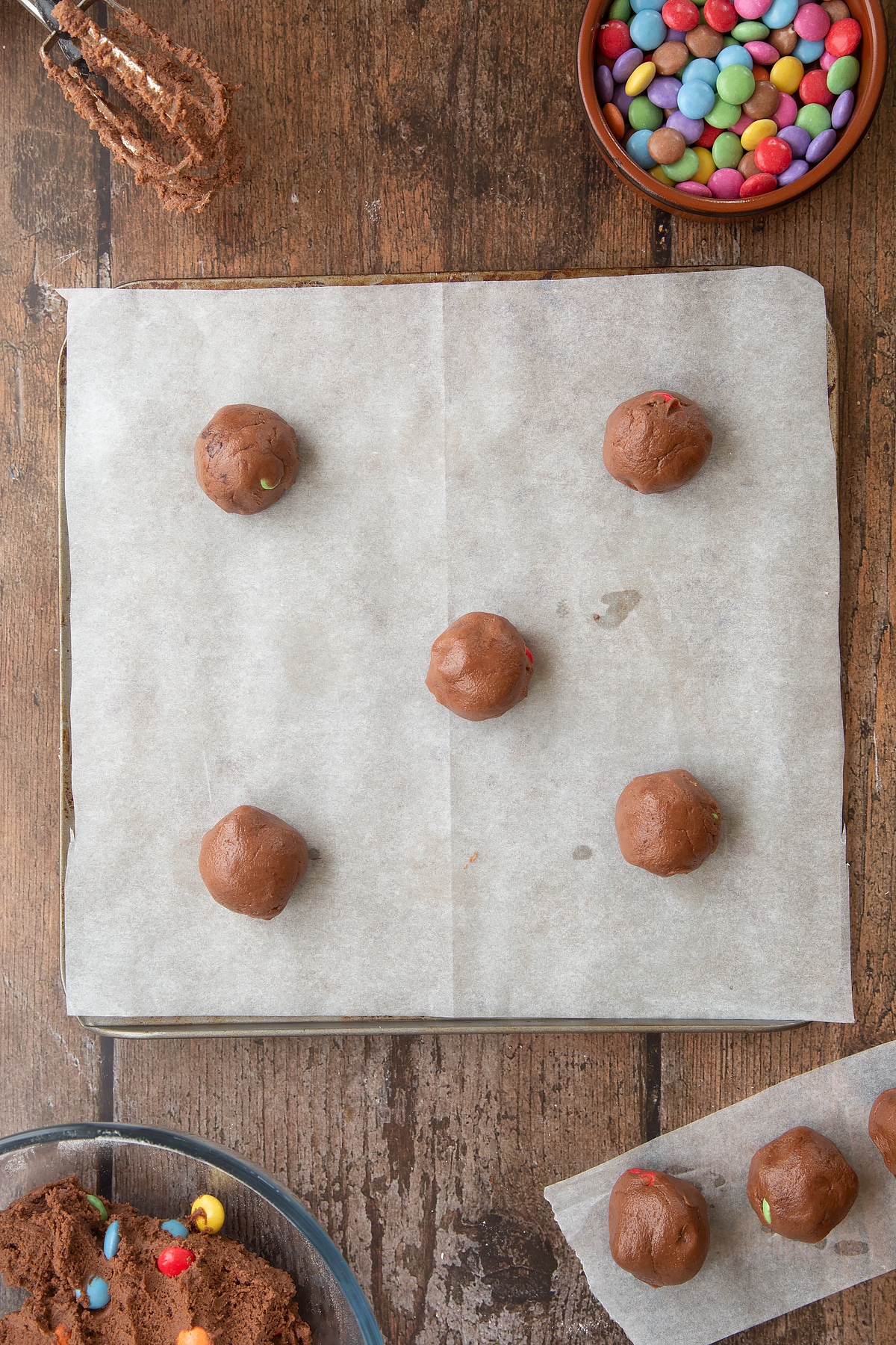 Balls of Smartie cookie dough on a baking tray lined with baking paper. Ingredients to make the Smartie cookie recipe surround the tray.