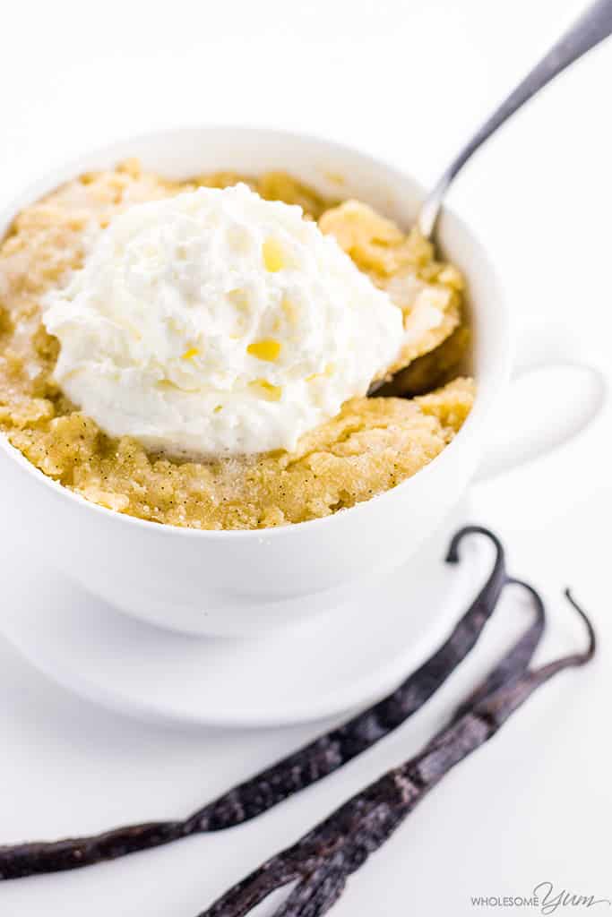 Keto paleo vanilla mug cake, topped with cream. A spoon delves into the cake. Two vanilla pods sit to the side of the mug.
