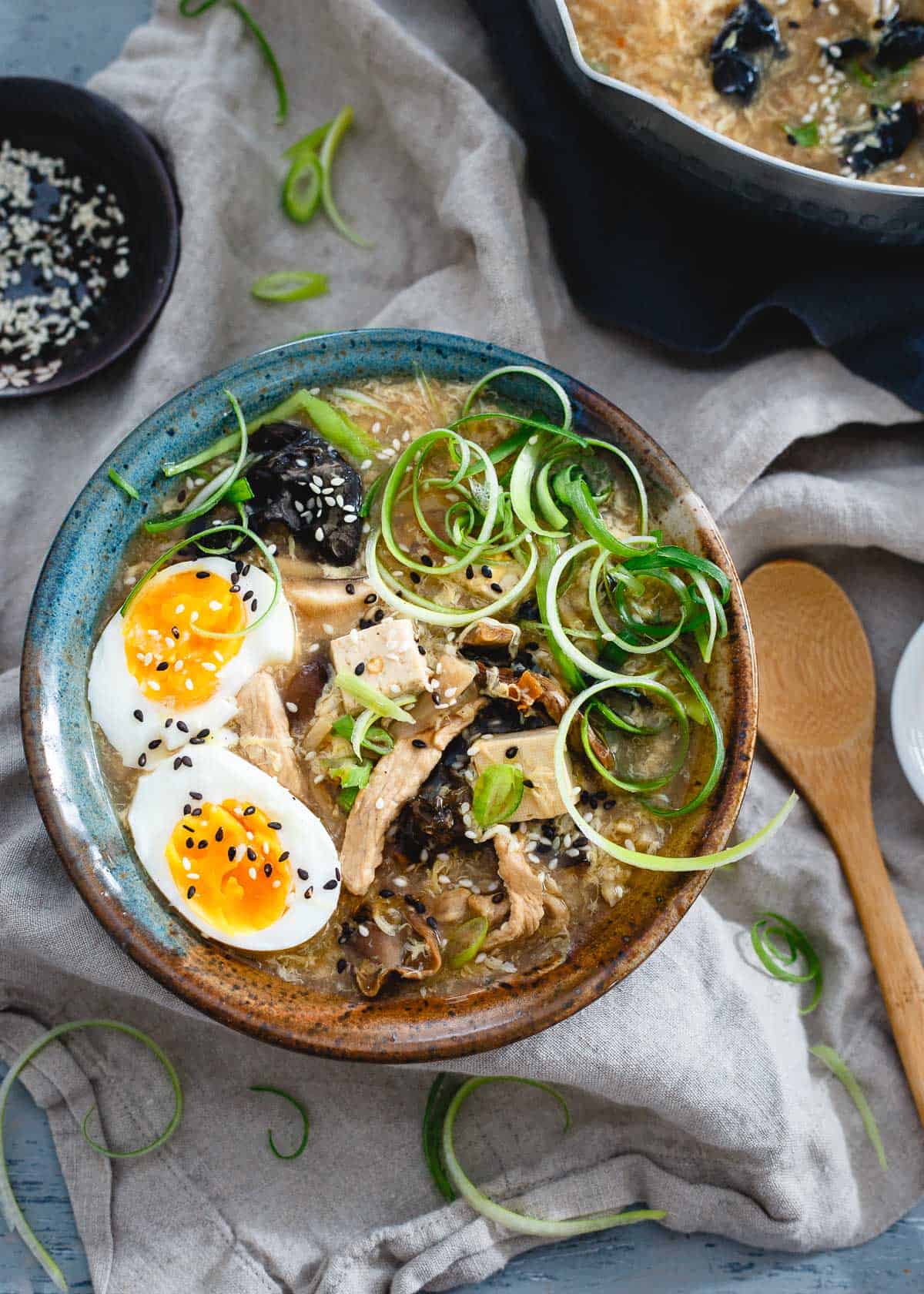 Bowl of hot and sour egg drop soup made from tofu on top of a table cloth with a bowl of sesame seeds at the side.