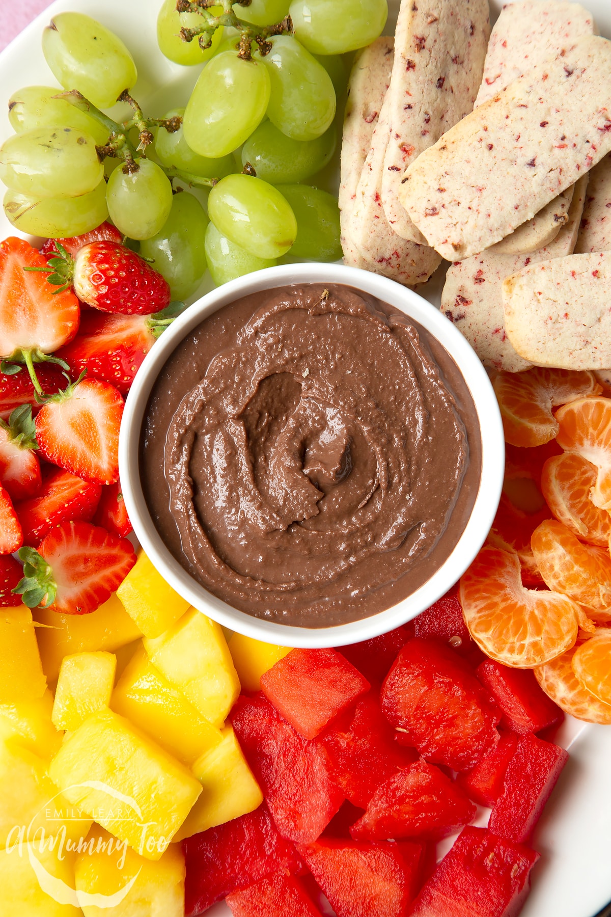 Strawberry biscuits surrounded different fruits. In the center there's a bowl of chocolate spread.