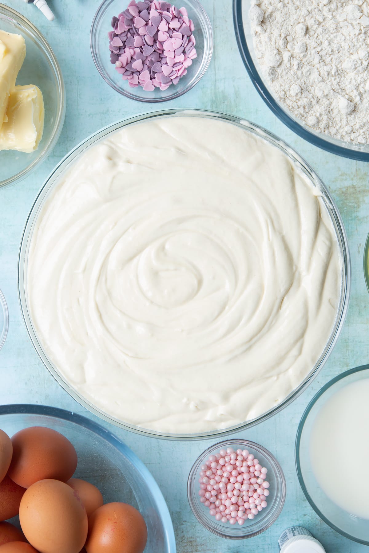 Pale cake batter in a mixing bowl. Ingredients to make pink ombre cake surround the bowl.