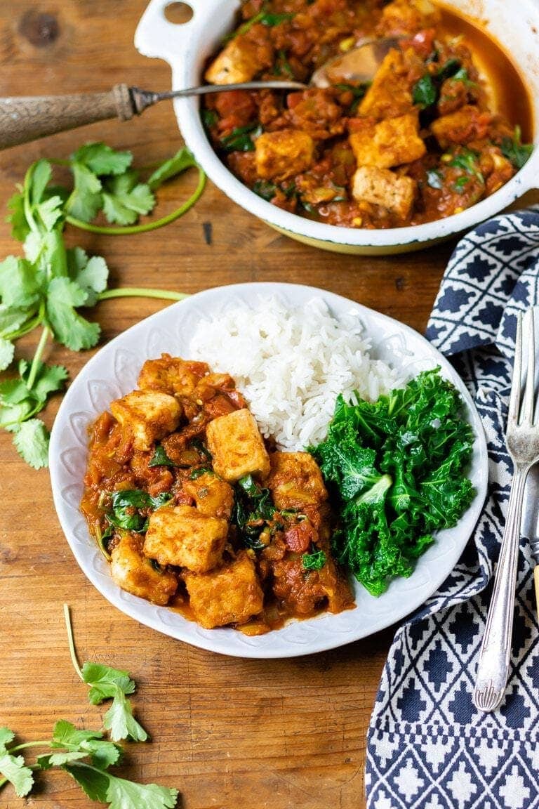 Tofu curry with a side of kale and white rice inside a deocrative plate. On the side there's a decorative blue and white napkin which holds a knife and fork.